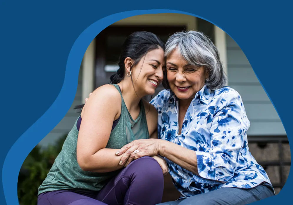 Two women sharing a warm, affectionate moment sitting together on a front porch. A younger woman in a green tank top and purple leggings leans close to an older woman with gray hair wearing a blue and white floral blouse. Both are smiling as they hold hands, suggesting a close family bond, likely mother and daughter. The photo is framed with an organic, blob-shaped border against a blue background.